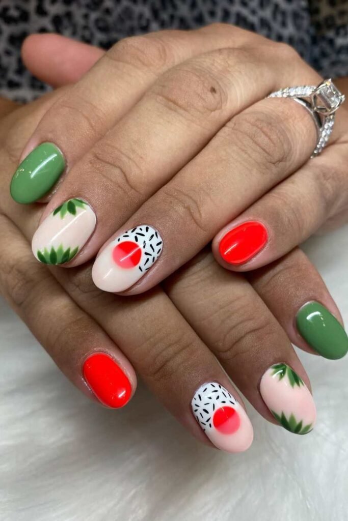 Short almond nails with a mix of green, red, and nude polish featuring leafy designs, red half-moons, and black-and-white sprinkle patterns—perfect for a playful spring break manicure.